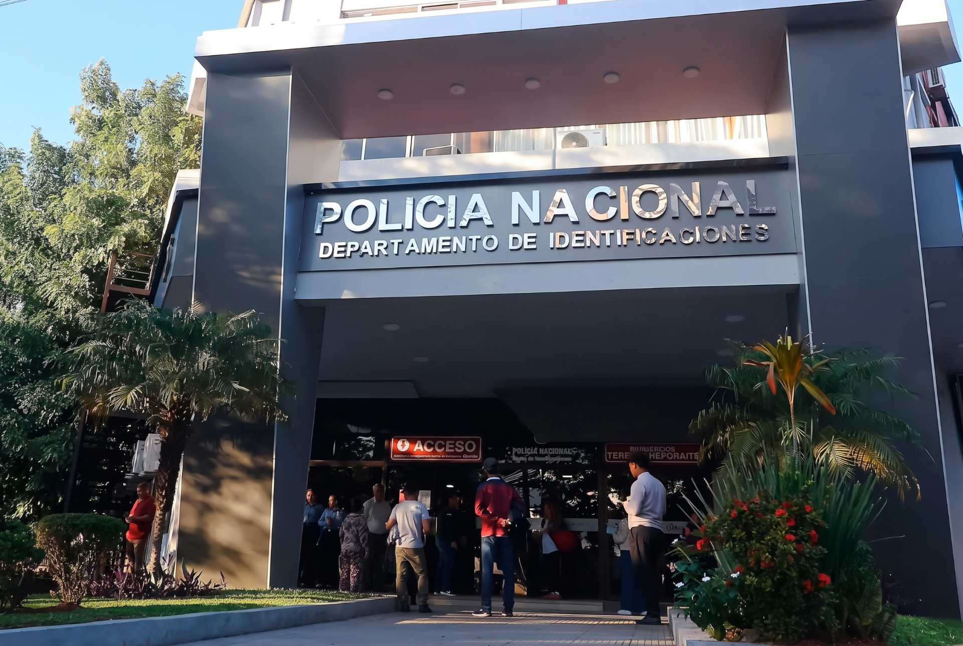 Eingangsbereich des Departamento de Identificaciones der Policía Nacional mit silberner Großbeschriftung über dem überdachten Portal, davor Wartende, Rasen, Palmen und rot blühende Pflanzen
